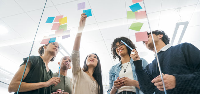 Five people looking at a glass wall as one is adding a post it note to a wall