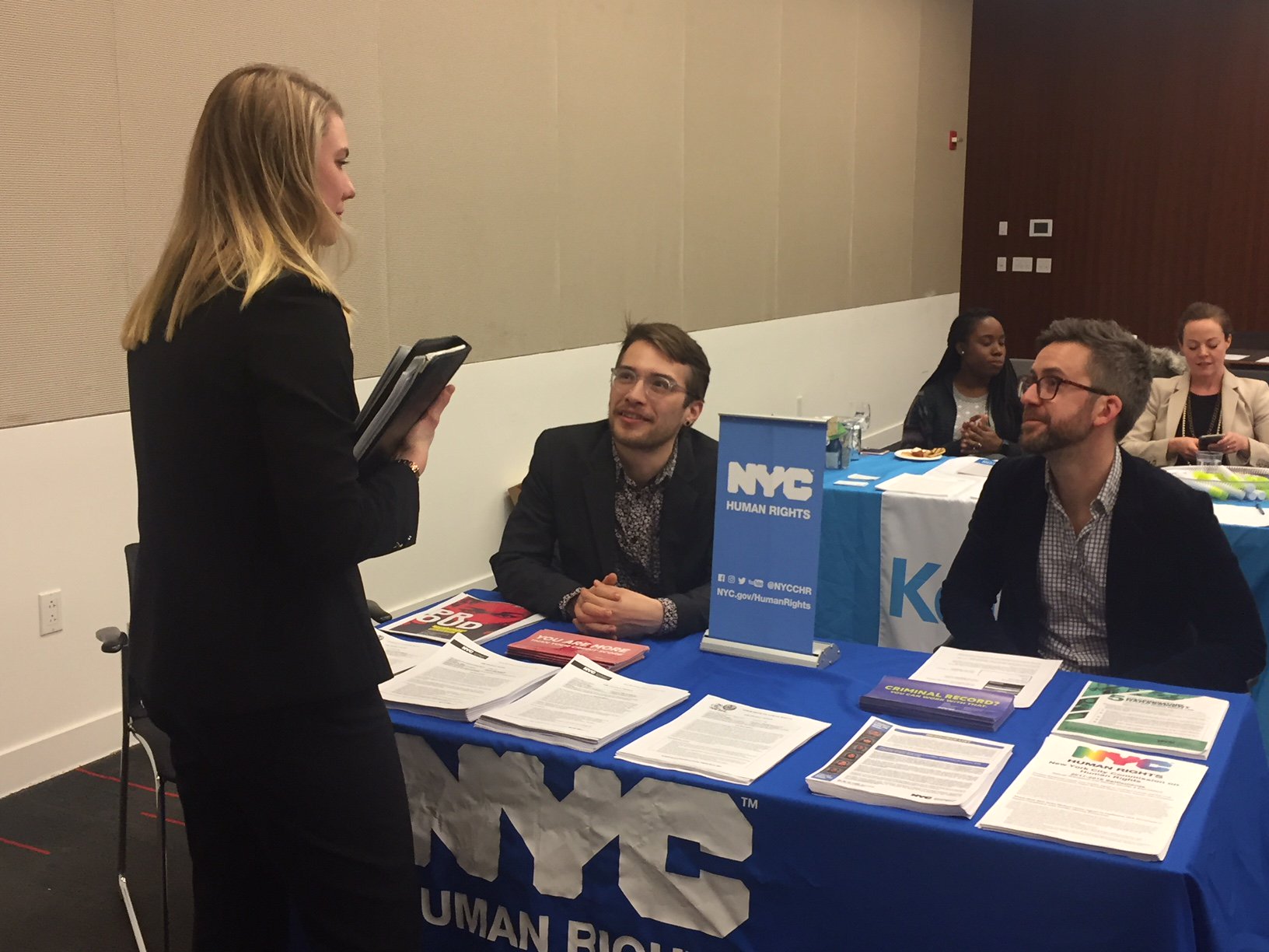 Two Commission staff members sitting at a table speaking with a woman.  Tablecloth says, “NYC Human Rights” and there are Commission materials on the table.