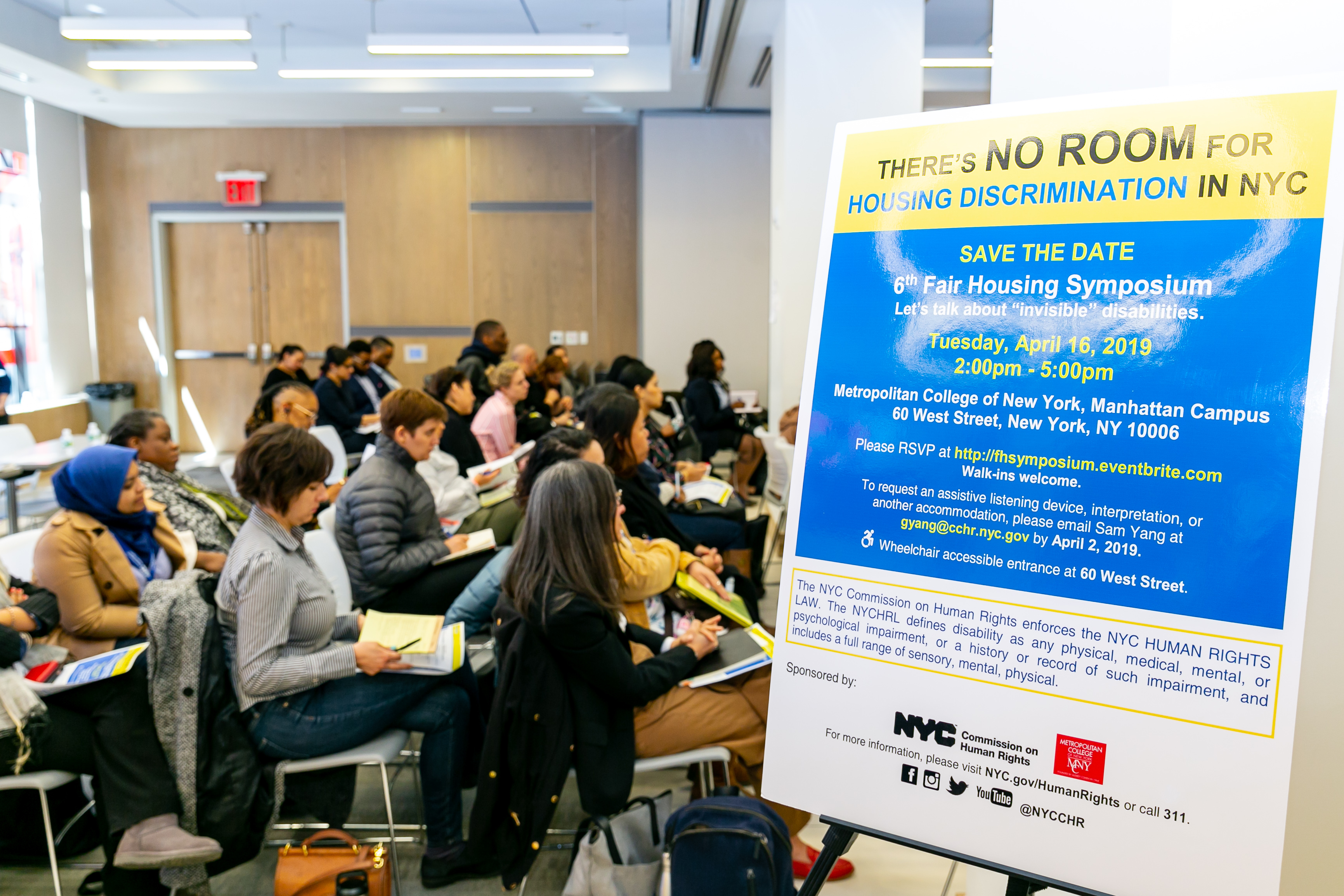 Profile of audience sitting at event next to a sign that reads There's No Room For Housing Discrimination in NYC, 6th Fair Housing Symposium, Tuesday, April 16, 2019.