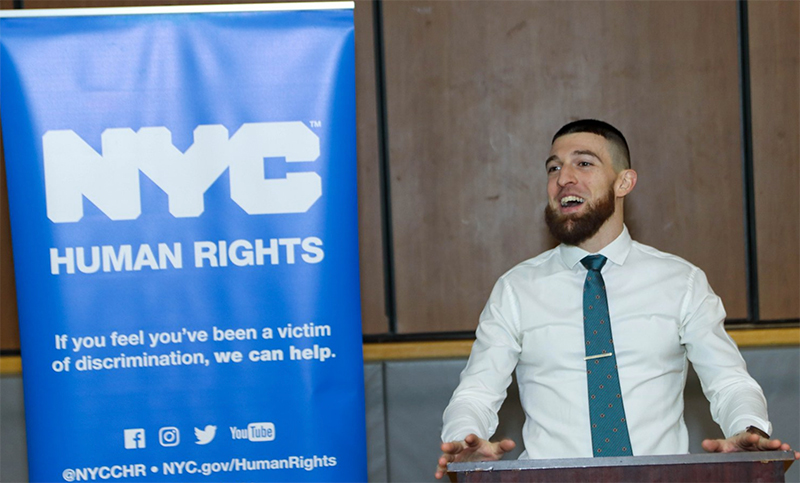 Commission employee speaking at a podium with blue banner behind him that reads in white lettering, "NYC Human Rights. If you feel you've been a victim of discrimination, we can help. @NYCCHR. NYC.gov/HumanRights."