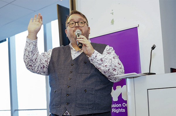 A man is presenting at an event with a microphone up to his mouth and his hand raised. NYC Commission on Human Rights banner is behind him.