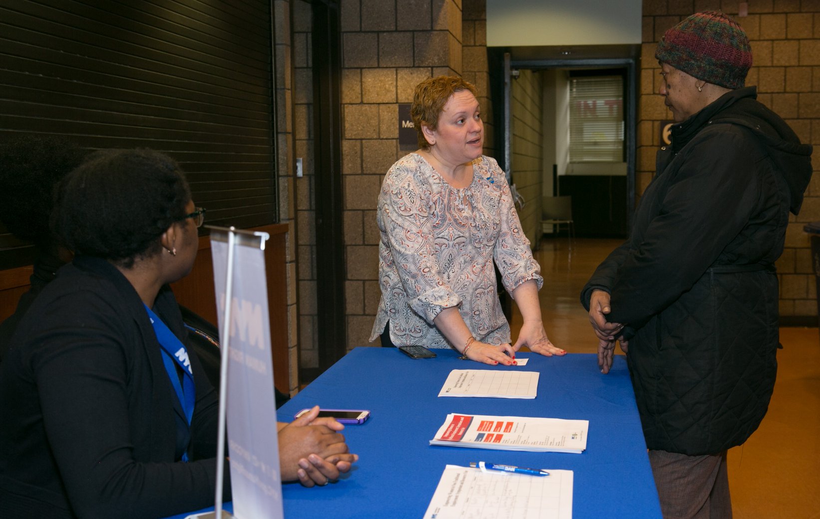 Two people (one Commission staff member and one member of the public) speaking at Commission table, while a seated staff member looks on.