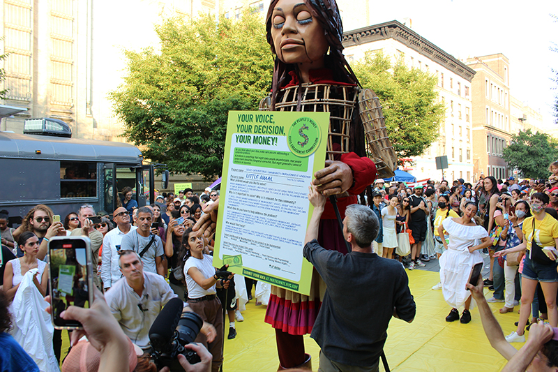 Photo of Little Amal holding a People's Money sign