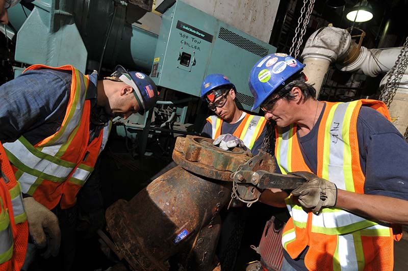 Three city staff installing a Y-strainer as part of a capital HVAC system upgrade. Three city staff installing a Y-strainer as part of a capital HVAC system upgrade.