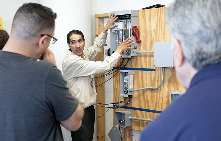 Instructor uses a sample inverter to teach Solar PV installation concepts to a group of students looking on.