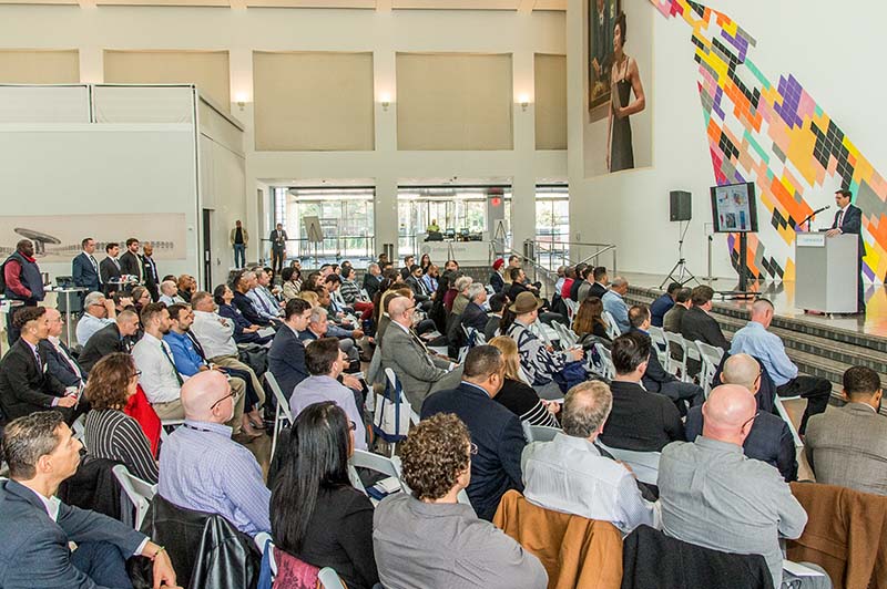 A lecture being conducted in a large open area with a stage and a diverse group of participants. On stage is the lecturer and a screen.  A lecture being conducted in a large open area with a stage and a diverse group of participants. On stage is the lecturer and a screen.