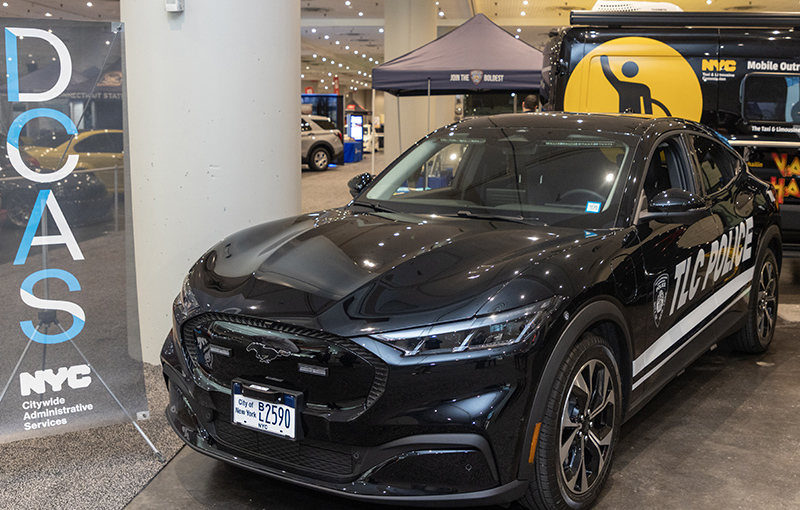 A Mach-E electric vehicle marked with 'TLC Police' displayed indoors at an NYC Citywide Administrative Services (DCAS) event, with a Mobile Outreach Unit van and promotional booths in the background.