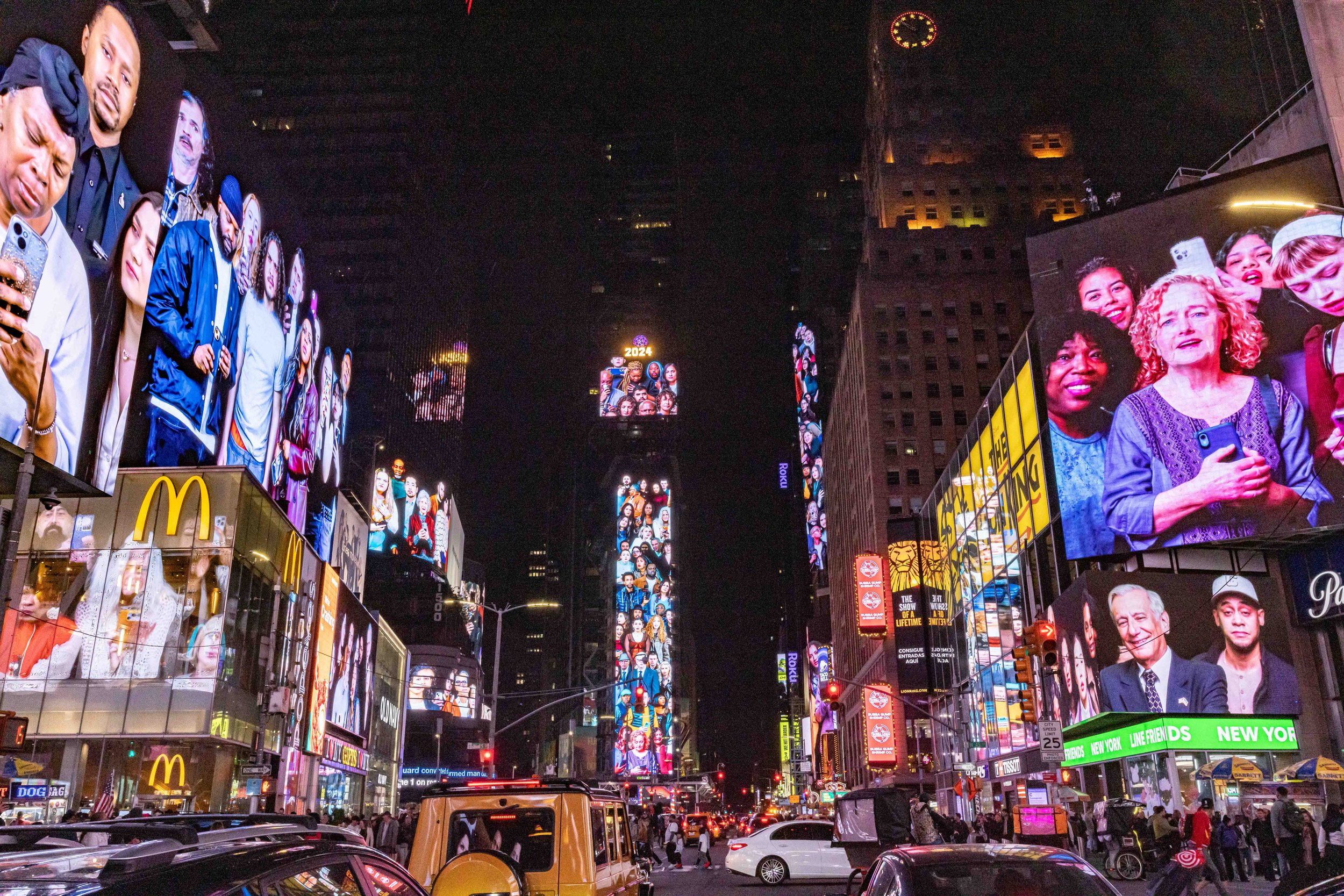 A photograph of Times Square at night shows images of diverse crowds facing the the streets across multiple large LED screens