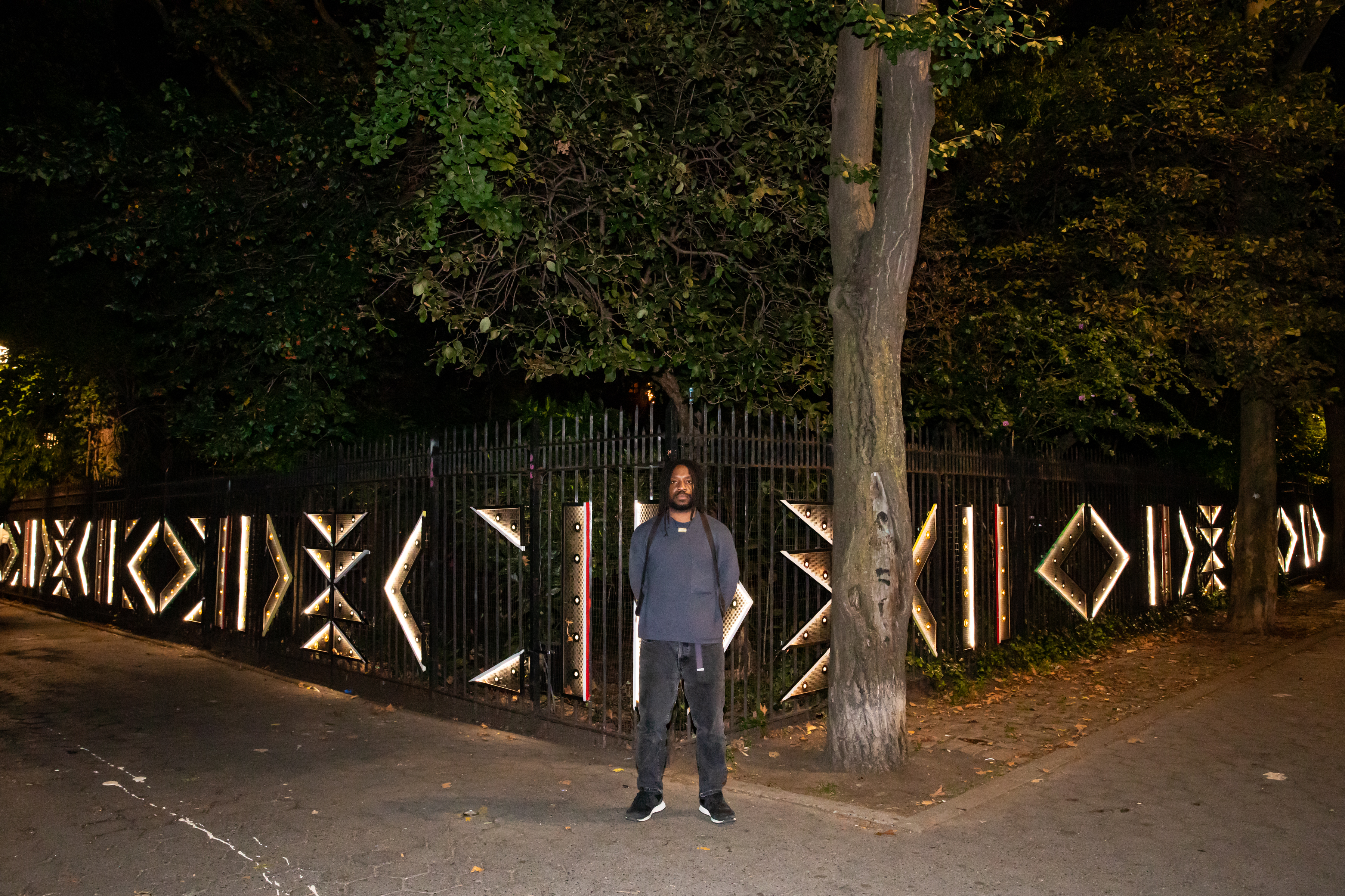 Photograph shot at night of Immanuel Oni standing in front of his illuminated installation mounted on the fence of M'finda Kalunga Garden