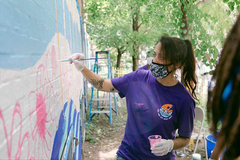 Artist wearing a mask and a City Artist Corps shirt paints a mural outside