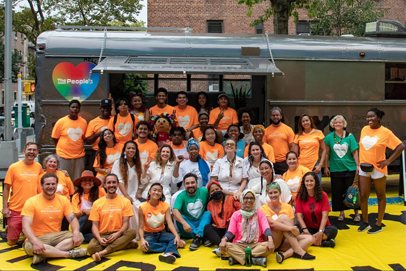 People sit in front of The People's Bus. Many wear orange People's Bus shirts.
