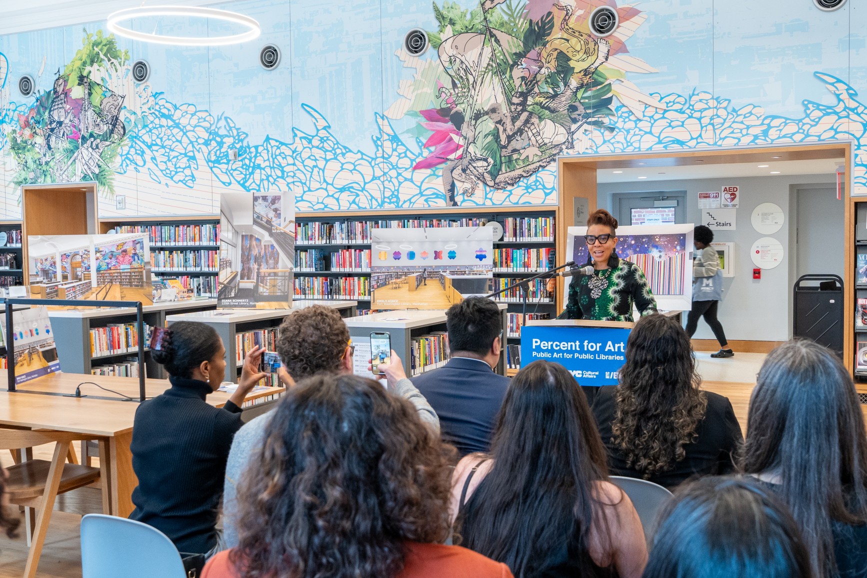 Commissioner Cumbo speaks at a podium with art by David Rios in the background.