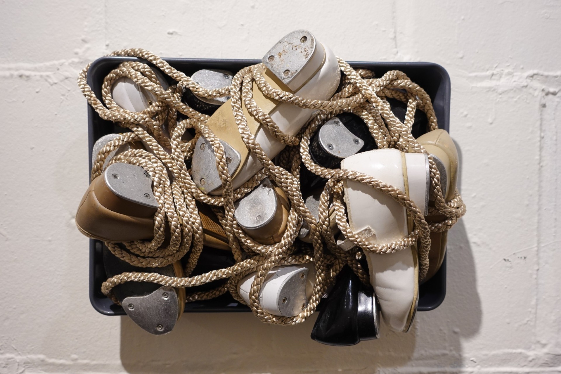 Small children's tap shoes are strung together with rope in a frame.