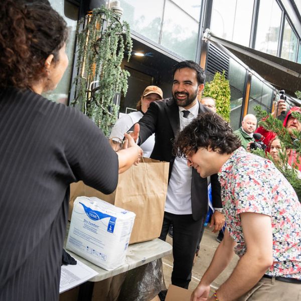 A man shakes a woman's hands outside next to a table with supplies while another man is reaching for the supplies outside of a restaurant.