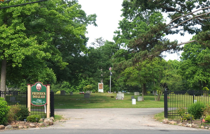Entrance to fenced cemetery with gravestones, memorial, and flagpole, sign reads 'Frederick Douglass Historic Memorial Park'
