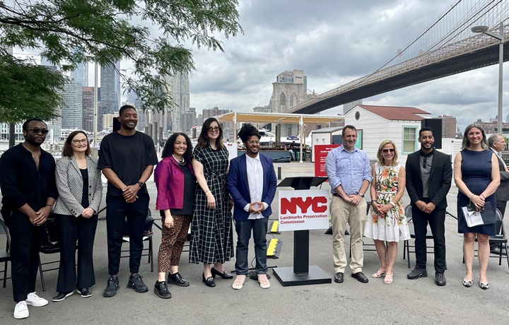 : Group of smiling people standing around podium with Brooklyn Bridge and NYC skyline in background 