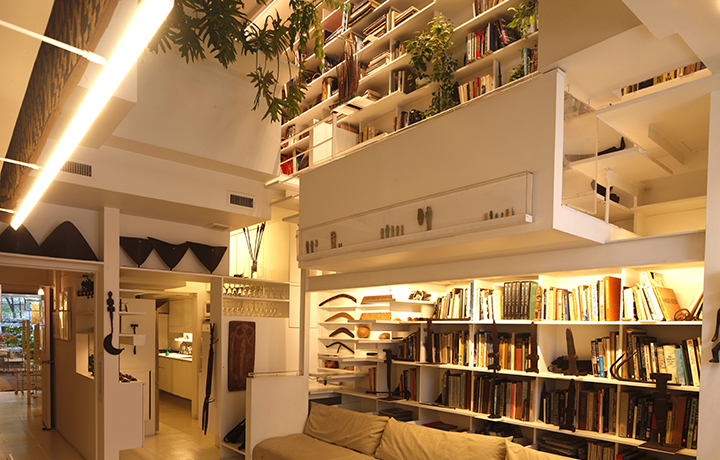 Image of apartment interior looking down from balcony to the floor below. Walls and furniture are largely white, as staircase on the right side of photo.  Large plant hanging over second floor balcony level.