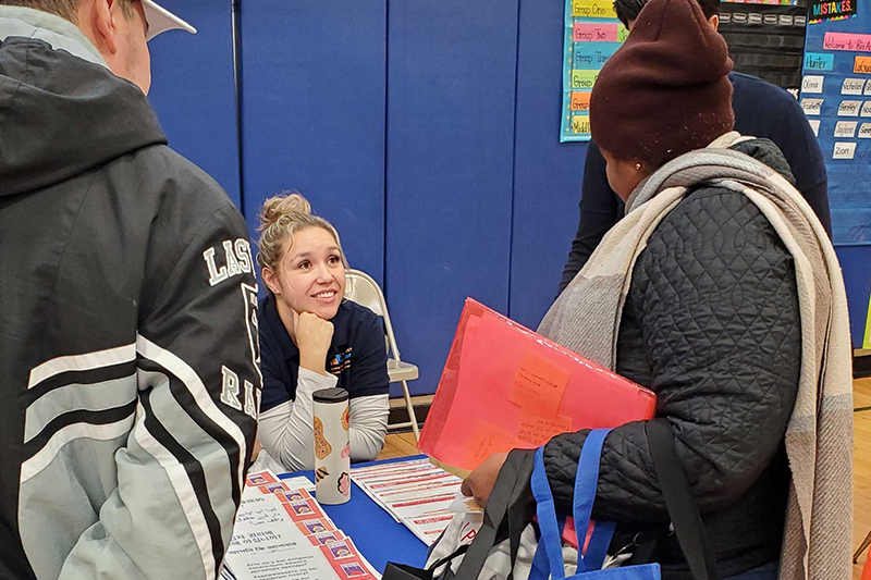 A P E U Specialist sits at a table and speaks with people who have stopped by for resources