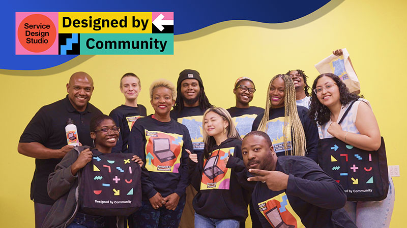 A group of community members and NYC staff pose for a picture with matching Designed By Community bags and sweaters. A Service Design Studio Designed by Community logo banner overlays the image.