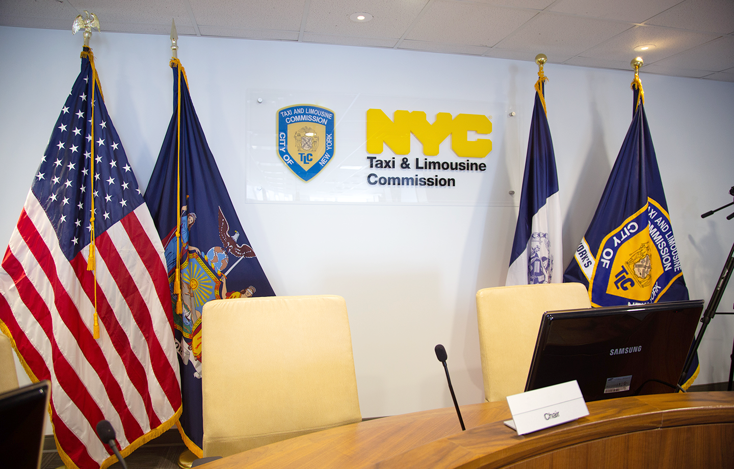 Conference room with a Taxi and limousine commission logo on the back wall, an American flag, a New York State flag, a New York City flag, TLC flag, two chairs behind a desk a table microphone and a computer screen. photo of a Commission meeting
