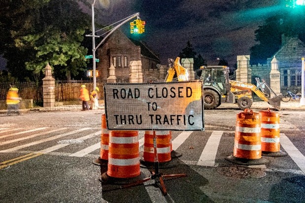 Orange barrels and a large sign reading “Road closed to thru traffic” stops traffic before roadway crews perform construction on a street at night.