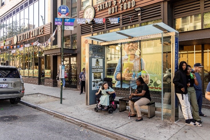People wait under and around a covered bus stop shelter in New York City. One person waiting sits in a wheelchairs and a second person sits on a bench while holding a white cane.