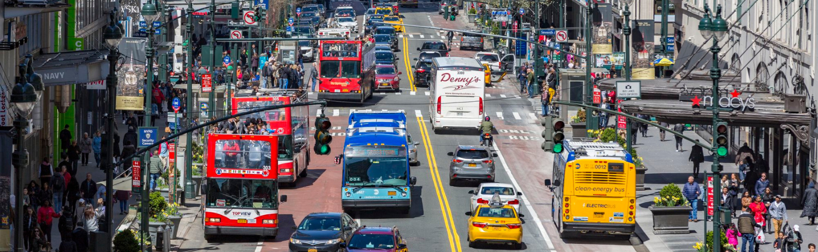 Red tour buses, blue city buses and other vehicles travel along a busy roadway in New York City