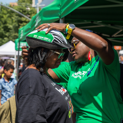 A woman wearing a green Vision Zero shirt fits a new bicycle helmet on another women's head at an outdoor event in New York City.