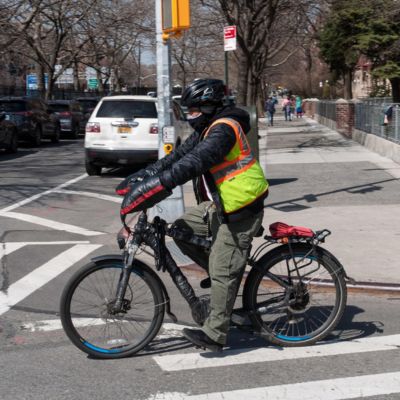 A delivery worker wearing a bike helmet and bright vest rides an e-bike in Brooklyn.