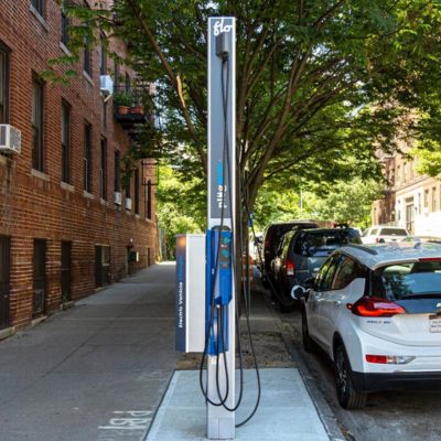An electric vehicle is parked on the curbside next to a gray and blue PlugNYC electric vehicle charging station on a N Y C sidewalk.