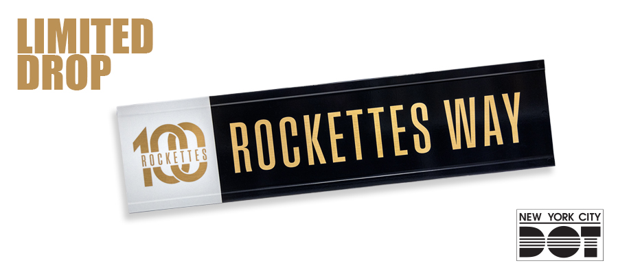  Black and white street sign with gold letters that say “Rockettes Way” with the number 100.