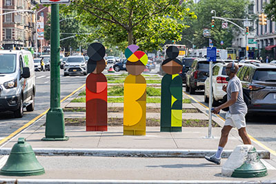 Three colorful statues representing women are installed on a concrete median along a busy roadway in Harlem. A man walks across the median in front of the sculptures. 