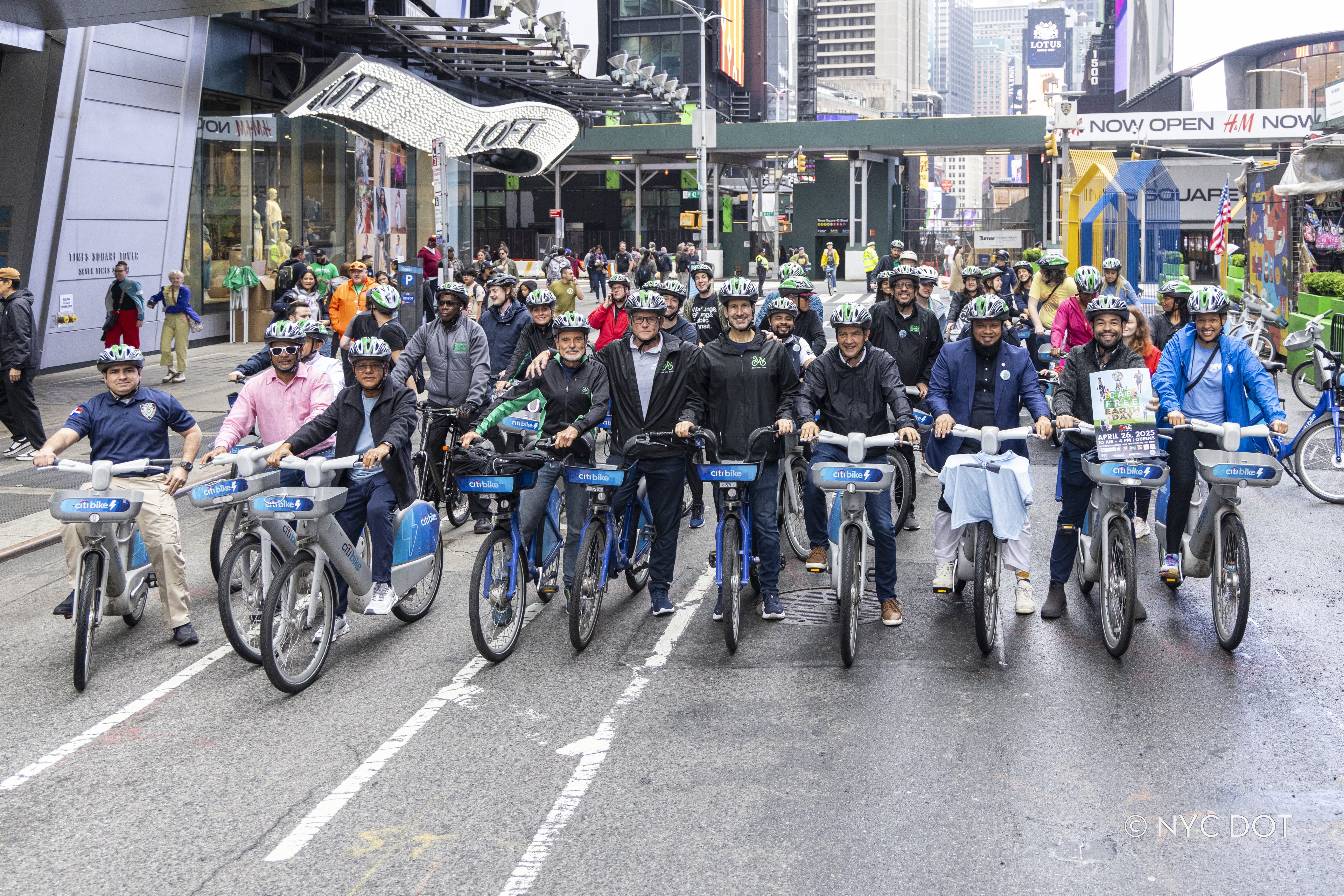 A large group of people wearing bike helmets stand or sit on bicycles and handcycles, ready to ride along a car-free roadway in Manhattan.