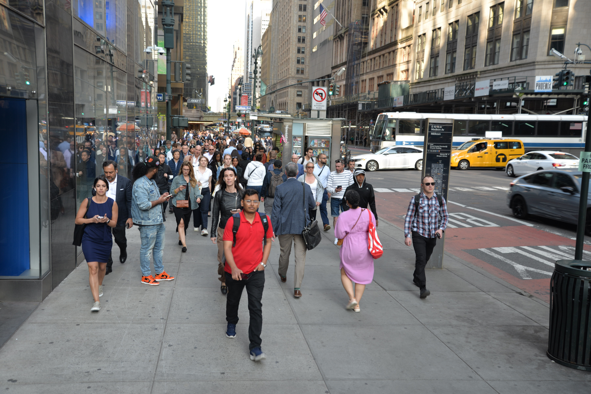 A large crowd of pedestrians walk in two directions along a wide sidewalk in Midtown Manhattan.