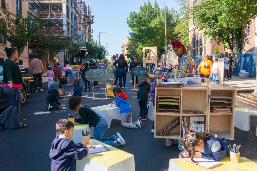 Students and teachers sit at pop-up tables and libraries on a street closed to vehicular traffic in front of a school.