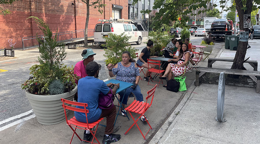 Multiple sit on red chairs placed on a pedestrian space next to the curb.